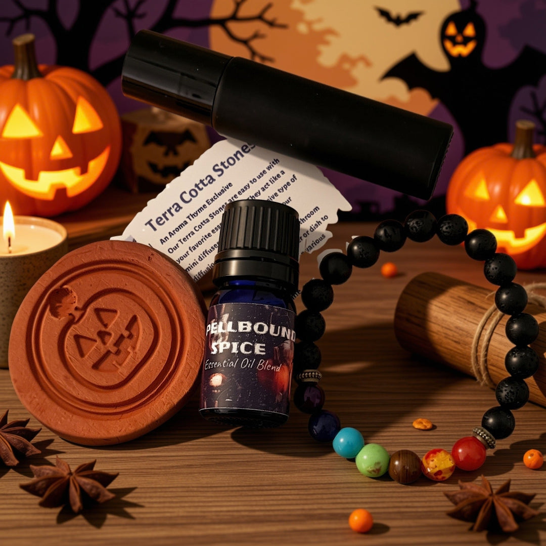 A Halloween-themed product arrangement on a wooden surface featuring a small bottle labeled 'Spellbound SPICE Essential Oil Blend,' a black cylindrical roll on container, a terracotta pumpkin diffuser, a beaded chakra bracelet, star anise, candy corn, and pumpkins in the background.